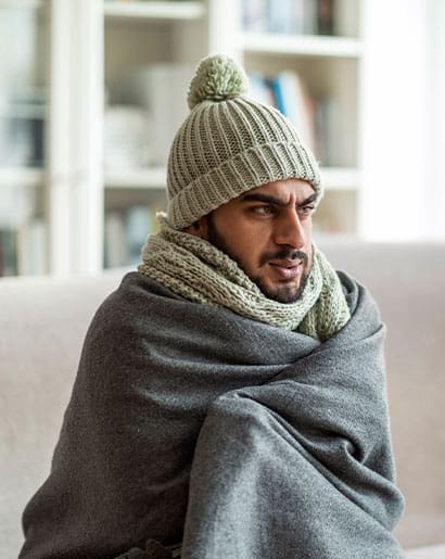 Man sitting on couch freezing with a hat and scarf wrapped in a blanket